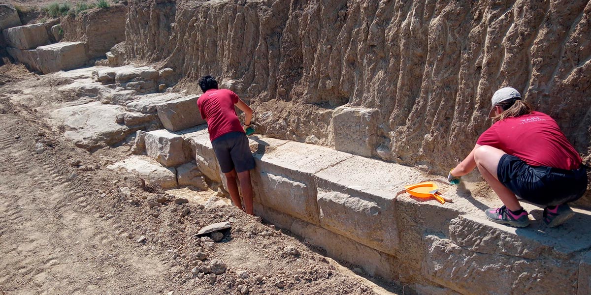 Dos estudiantes limpian los restos de lo que sería la muralla que cerraba la ciudad romana de Los Bañales 1